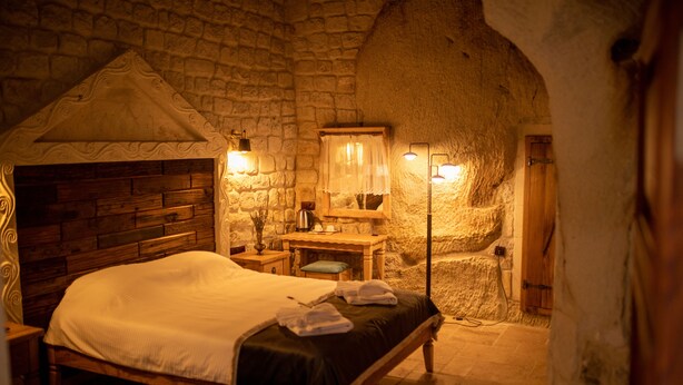 A stone-walled bedroom with a wooden bed, a nightstand, a desk, and a lamp. - Pome Granate Cave Hotel (Nevsehir)