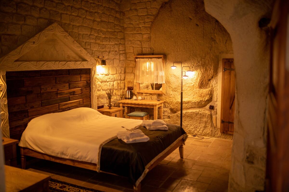 A stone-walled bedroom with a wooden bed, a nightstand, a desk, and a lamp.