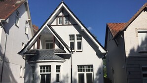 A two-story house with a steeply pitched roof, dormer windows, and a visible chimney. There are two cars parked in front. - Apartment am Wall (Emden)