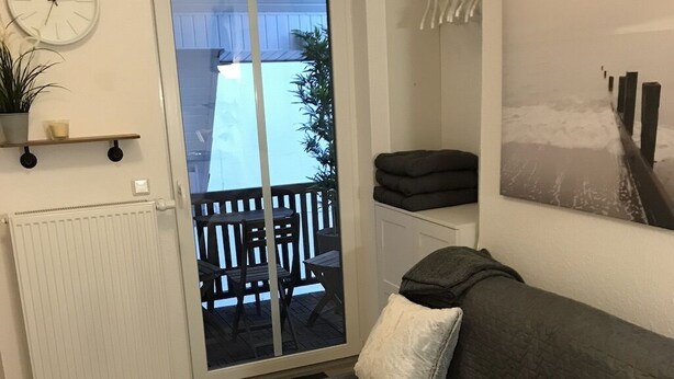 A living room with a grey sofa, white cushions, a small table with vases, a clock, and a sliding glass door leading to an outdoor patio with a table and chairs. - Apartment am Wall (Emden)