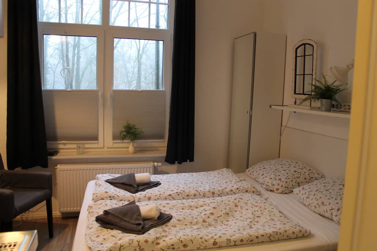 Ein Zimmer mit zwei Betten, einem Sessel, einem Heizkörper, einem Fenster mit Vorhängen, einem Spiegel und einem Regal mit einer Pflanze.