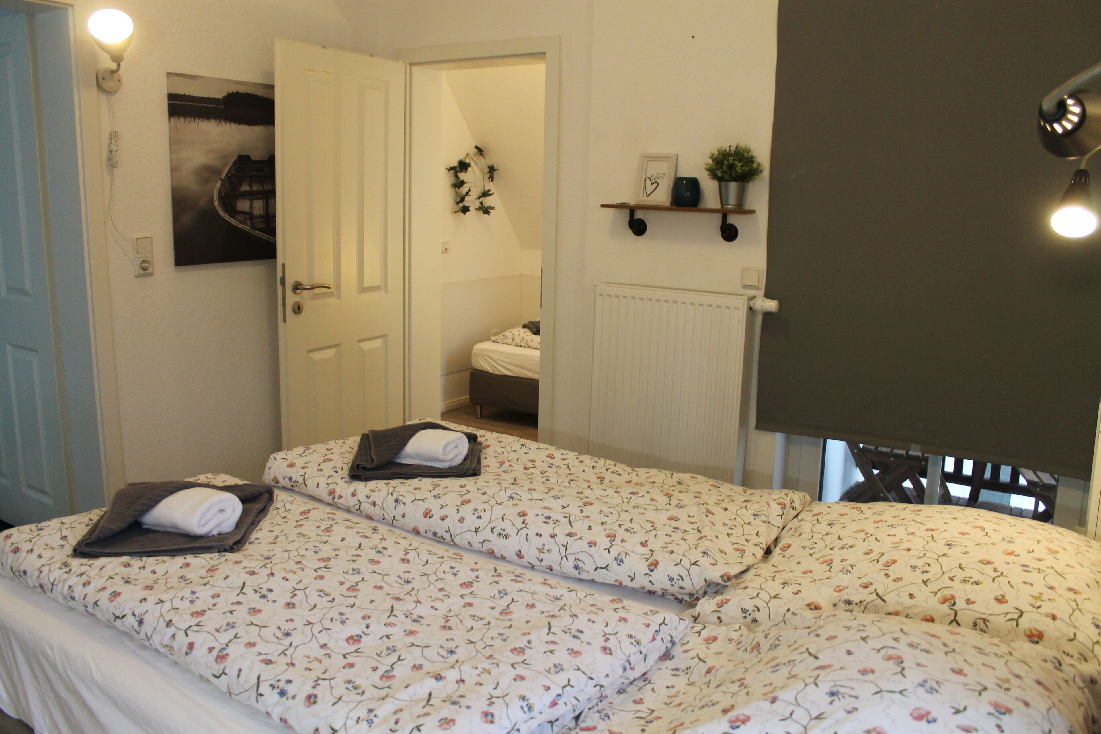 A bedroom with two beds, a radiator, a shelf with a plant, and a framed picture on the wall.