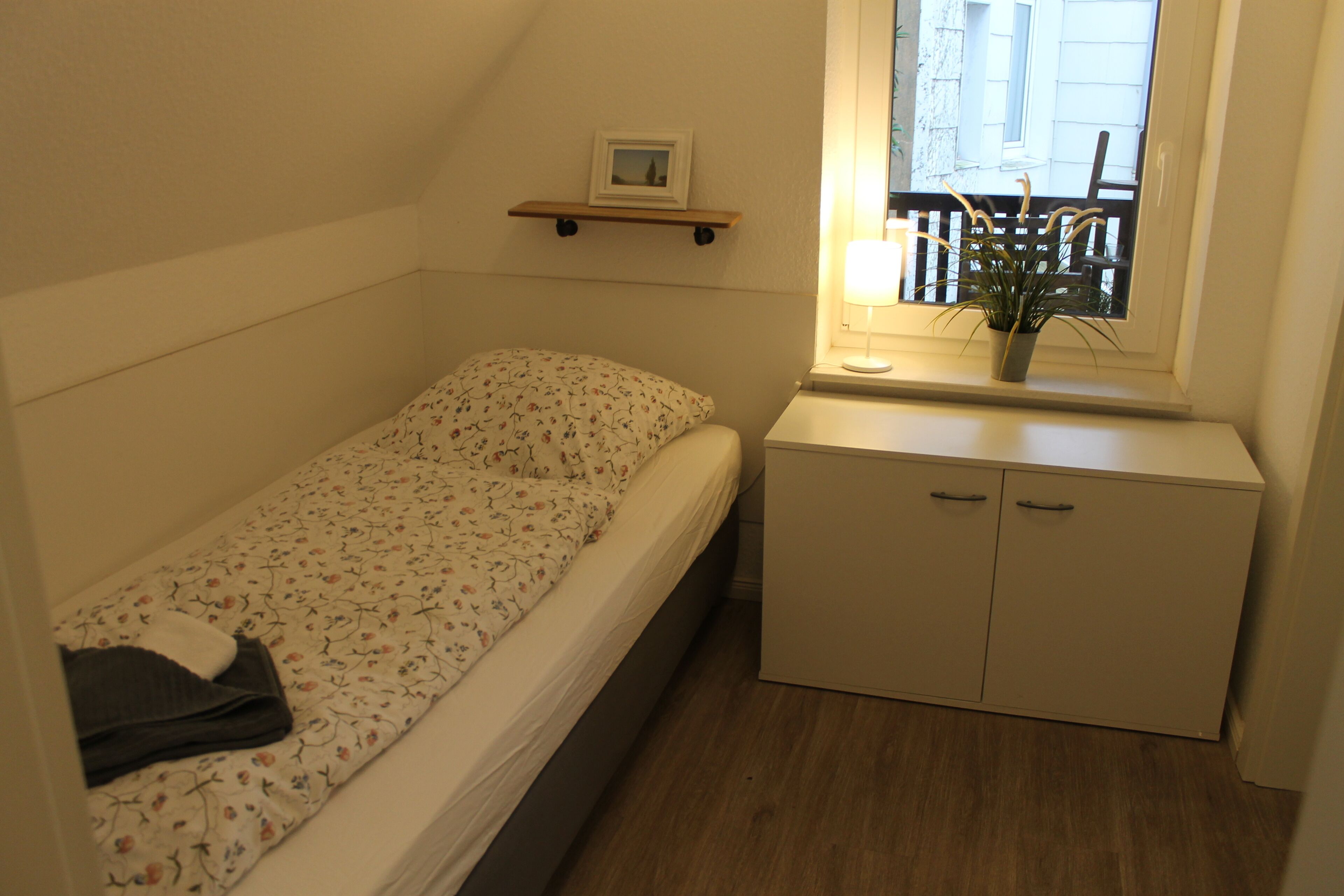 A small, single-bed room with a wooden headboard, a bedside table with a lamp, a small shelf with a picture, and a window with a view of a balcony.
