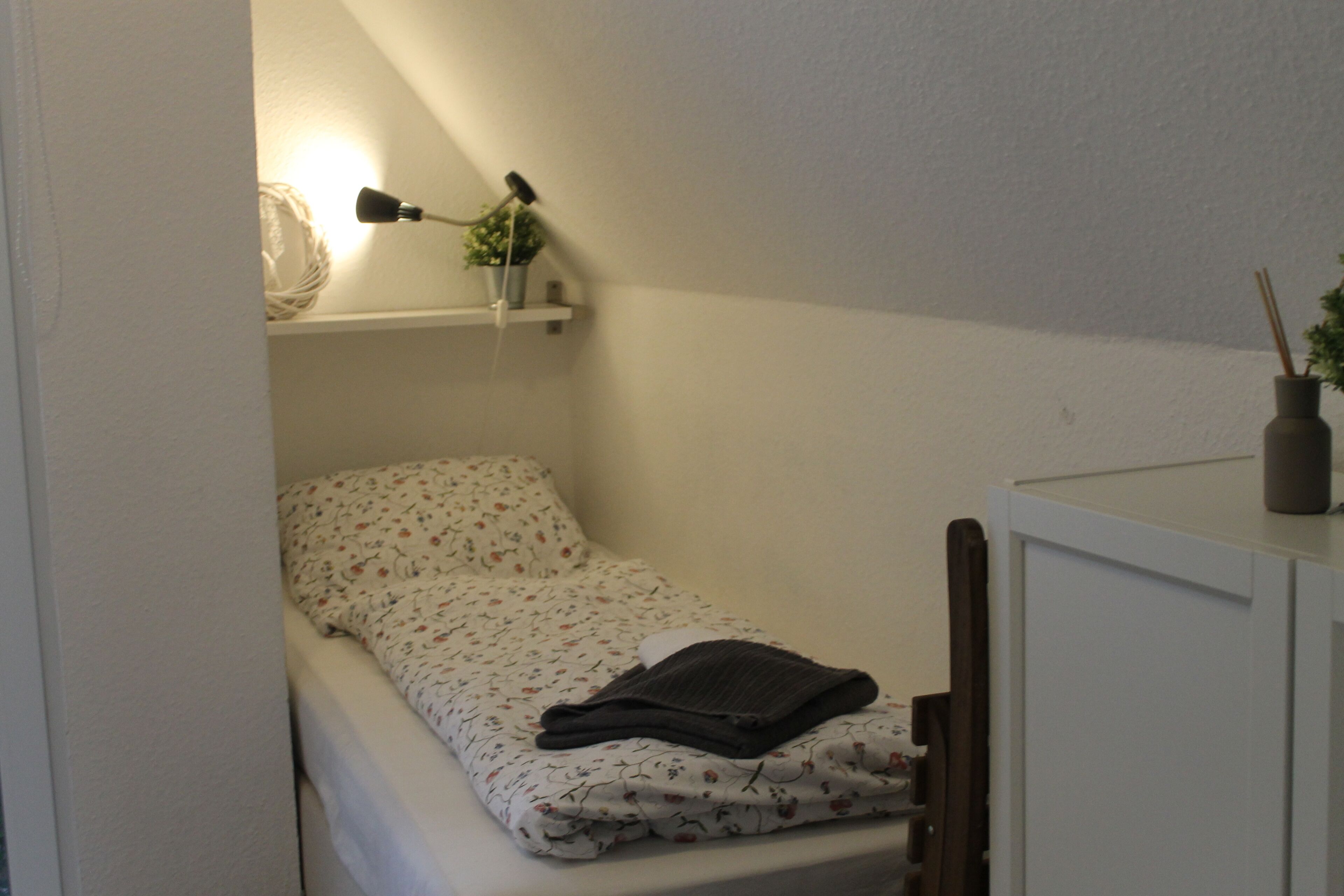 A small, neatly made bed with a floral-patterned bedspread and a folded dark-colored blanket. There is a shelf above the bed holding a lamp, a small plant, and a decorative item. A cabinet is visible to the right of the bed.