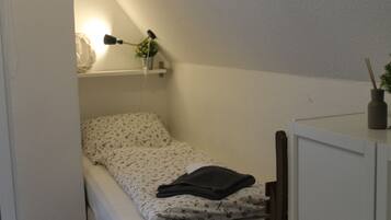 Een klein, keurig opgemaakt bed met een bedspreide met bloemendesign en een donkergekleurde, opgevouwen deken. Daarboven bevindt zich een plank met een lamp, een klein plantje en een decoratief object. Aan de rechterkant van het bed is een kastje te zien.
