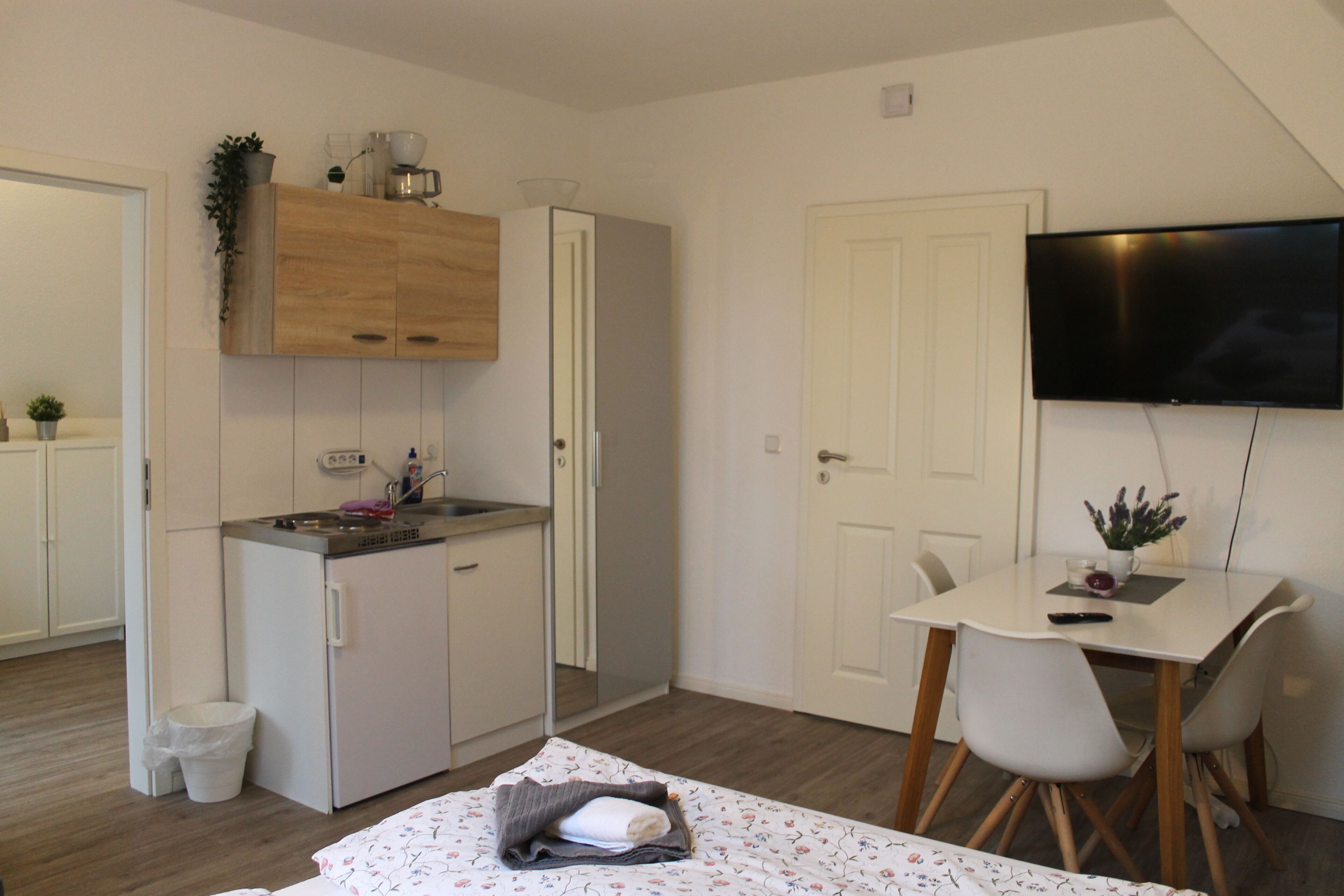 Een compacte kamer met een kitchenette, een bed met een keurig opgevouwen dekbed, een eettafel met stoelen en een aan de muur bevestigde televisie.