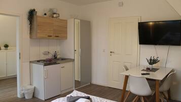 Een compacte kamer met een kitchenette, een bed met een keurig opgevouwen dekbed, een eettafel met stoelen en een aan de muur bevestigde televisie.