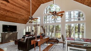 Un salón comedor espacioso con techo alto, vigas de madera, un televisor grande, chimenea y una zona de comedor con mesa y sillas.