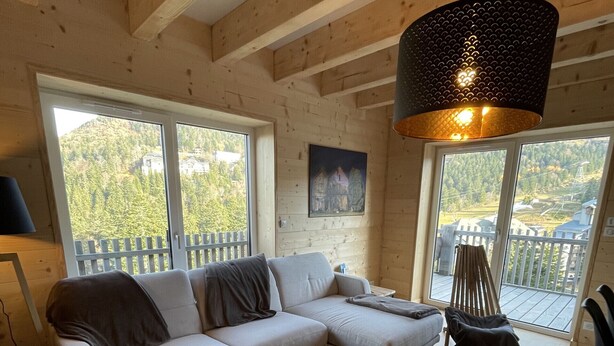 A cozy cabin interior with a wooden ceiling, a sofa, a coffee table, and large windows offering a view of a forested area. - Triplex - RDC les Ecrins (Le Lioran)