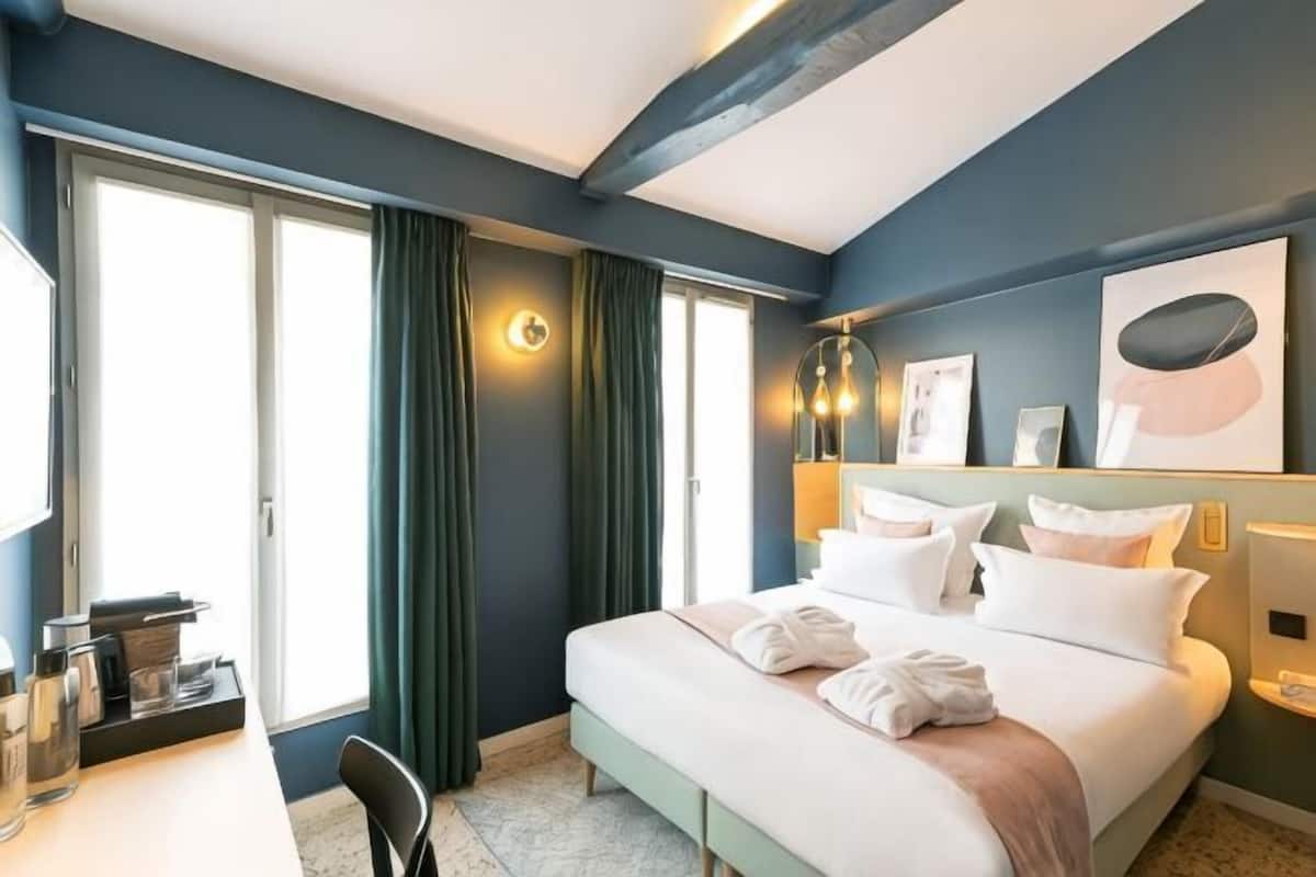 A hotel room with a bed, a desk, a chair, a large window with curtains, and artwork on the wall.