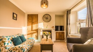 Ein Wohnzimmer mit einer Couch, einem Sessel und einem Tisch mit Blumen in einer Vase. Ein Kamin, eine Wanduhr und ein Fernseher auf einem Schrank sind ebenfalls zu sehen.