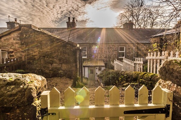 A stone building with a white picket fence in front, a stone wall, and a view of the sky with clouds. - Tring-on-Tor (Matlock)