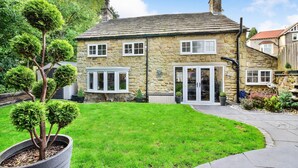 A stone house with a well-maintained lawn, a large potted tree, and a clear sky.
