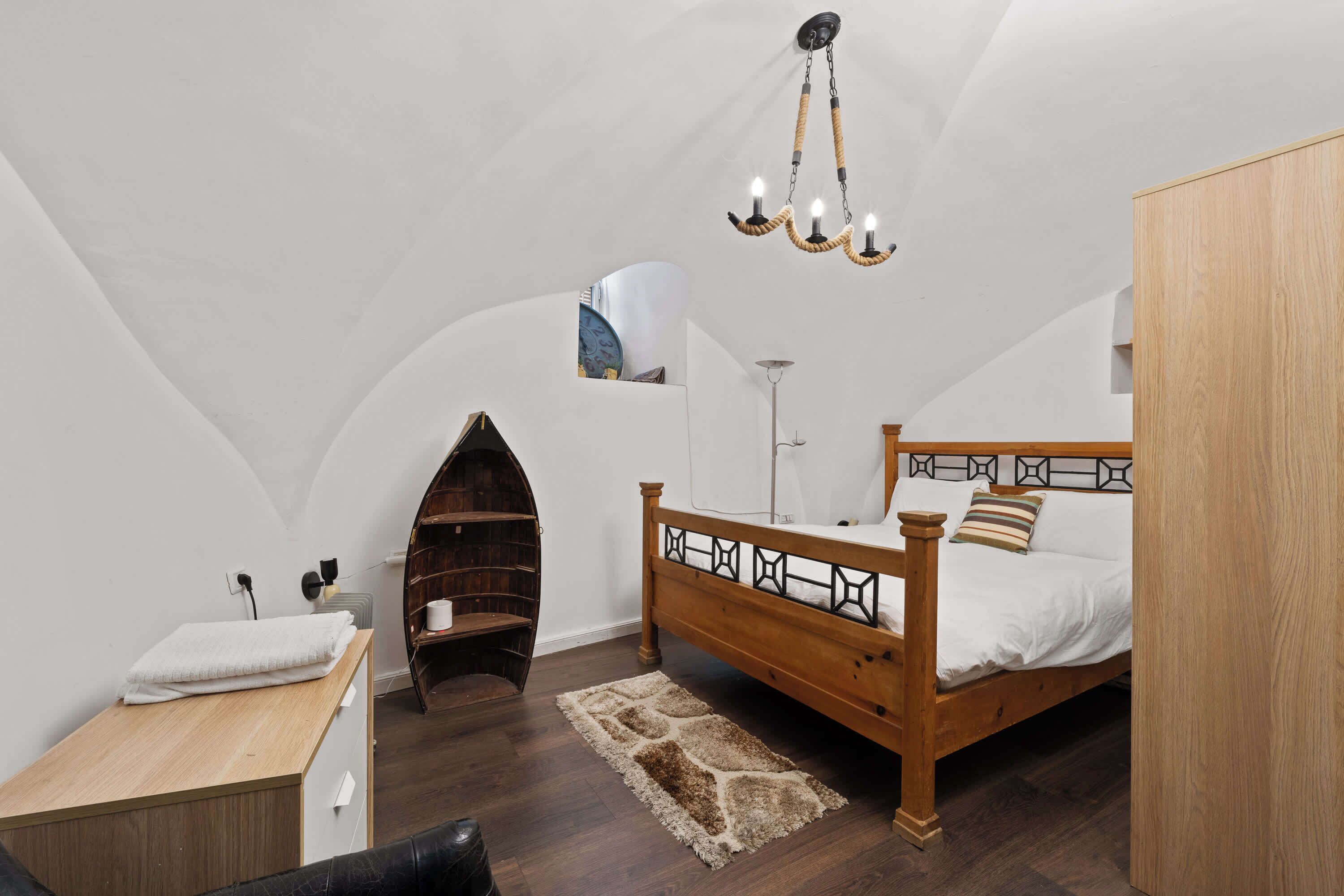 A bedroom with a wooden bed, a small desk, a chair, a wooden wardrobe, and a unique arched ceiling.
