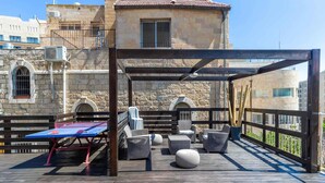 A rooftop terrace with a ping pong table, wicker furniture, and a stone building in the background.