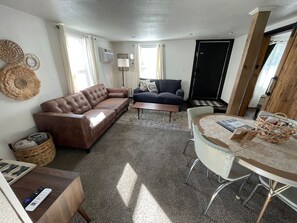 A living room with a brown leather sofa, a dark blue couch, a coffee table, and a round table with chairs. There is a wicker basket on the table and a remote control on the sofa. - N 7th Street · Fun Stay in the Heart of Rapid City (Rapid City)