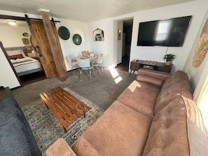 A living room with a large brown sofa, a wooden coffee table, a TV mounted on the wall, and a dining area with a white table and chairs.