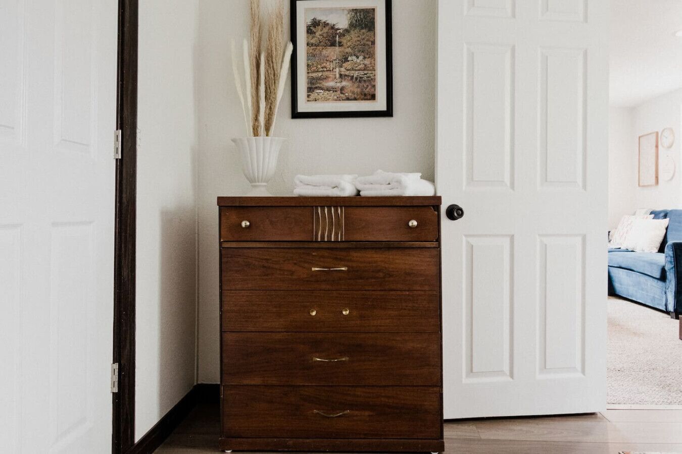 A wooden dresser with drawers and a small shelf, a framed picture on the wall, and a vase with dried plants.