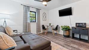 A modern living room with a sofa, a dining table with chairs, a ceiling fan, a flat-screen TV, and a microwave on a cabinet.