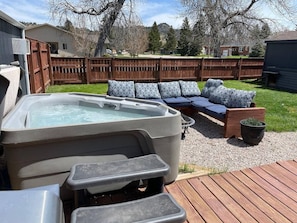 A backyard with a hot tub, a wooden deck, and a seating area with cushions.