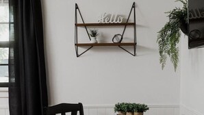 Un comedor con una mesa negra, dos sillas, un cartel que dice 'hello', un estante pequeño con una planta, y una maceta en la pared.