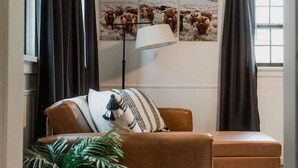 Un salón con un sofá marrón, una mesita auxiliar con una planta, una lámpara y un cuadro de vacas colgado en la pared.
