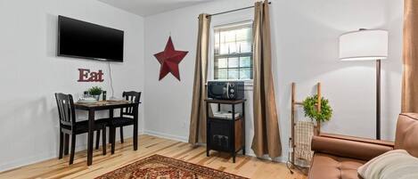 A compact living space with a dining area, a television mounted on the wall, a star decoration, a window with curtains, a sofa, and a floor lamp.