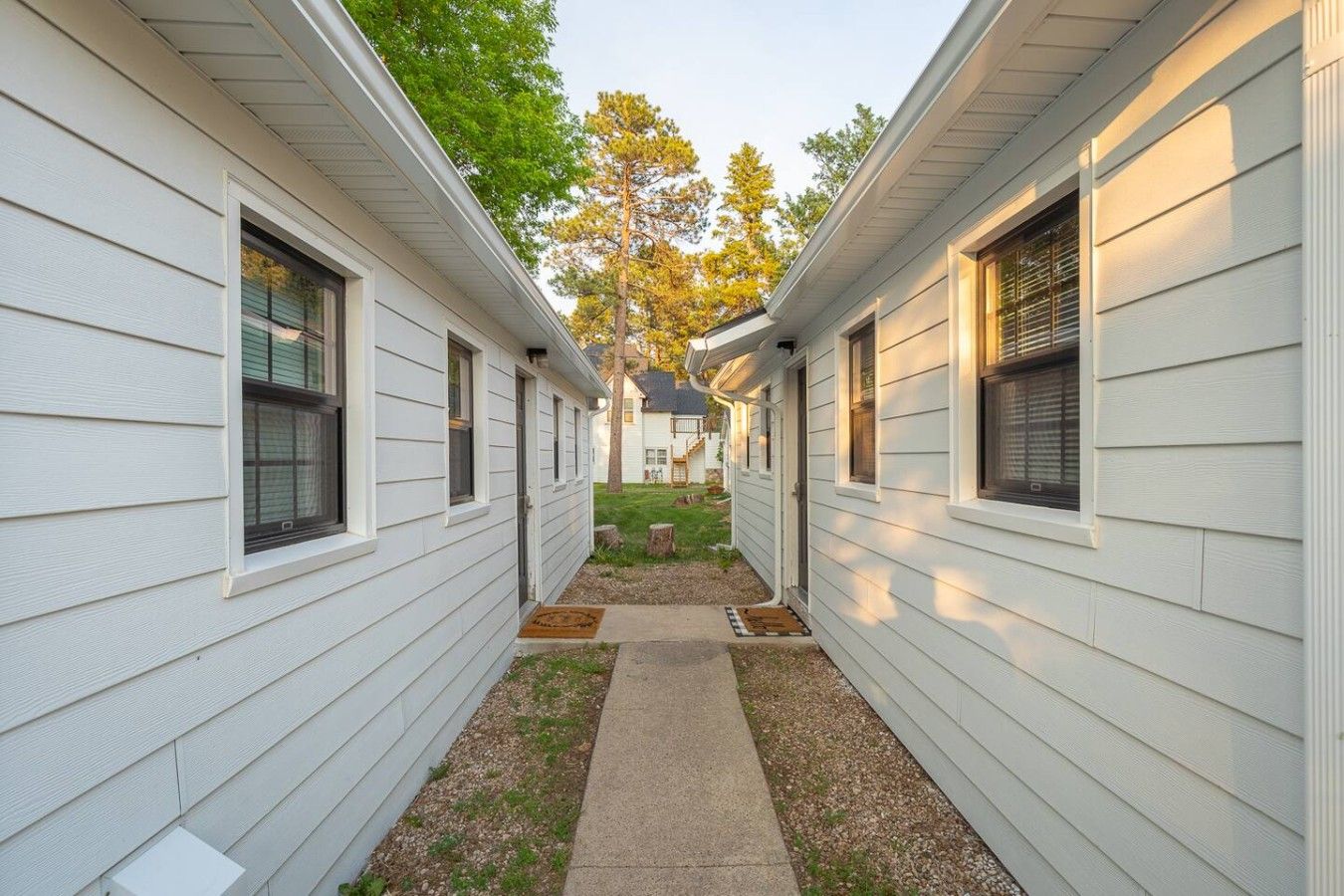 A narrow pathway between two white mobile homes with a grassy area and a small tree in the background.