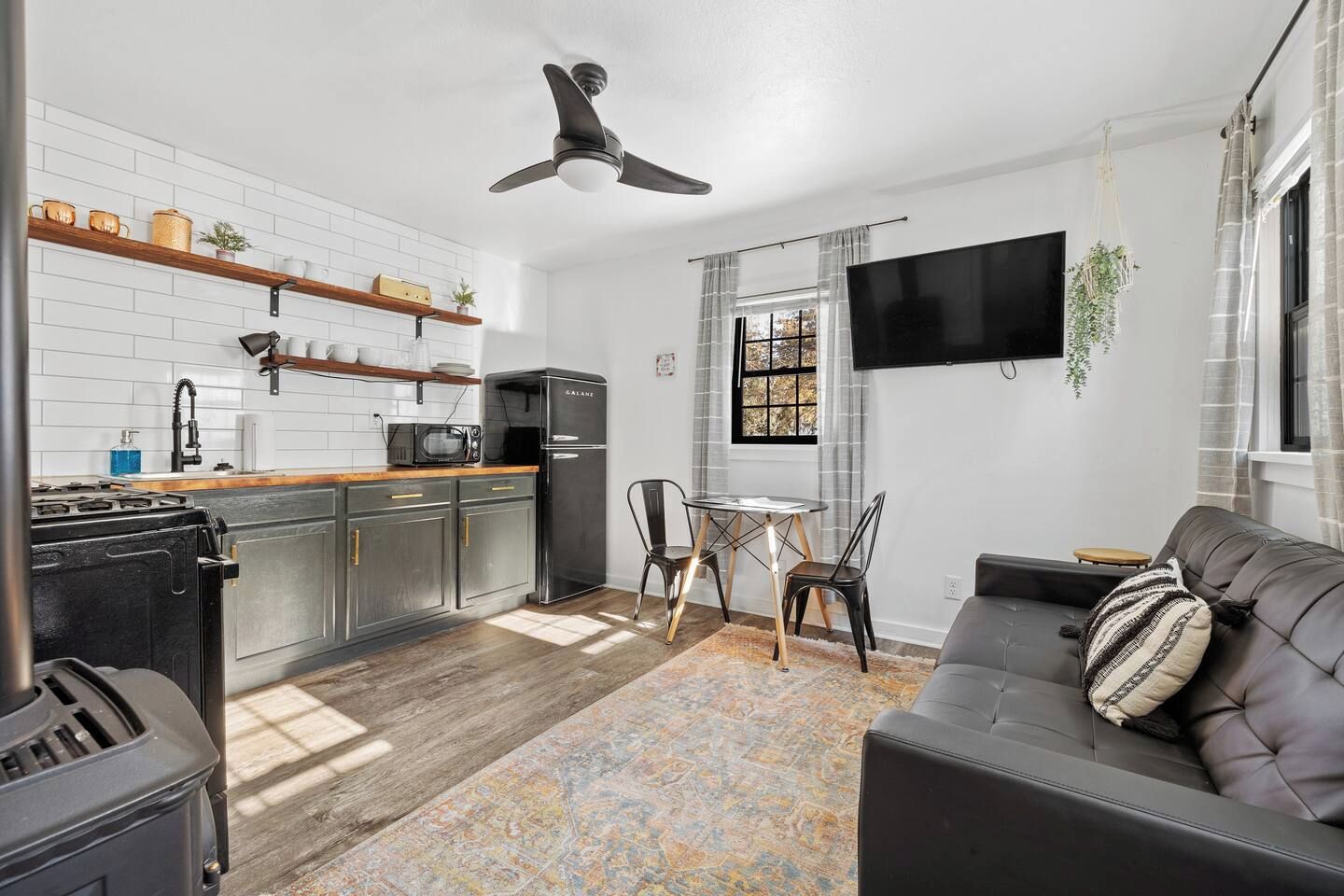 A modern kitchen with a black refrigerator, a microwave, and a stove. There is a dining area with a table and chairs, and a black leather sofa with a patterned pillow.