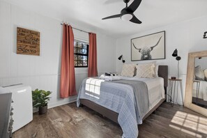 A bedroom with a bed, a ceiling fan, a window with curtains, a bedside lamp, a mirror, and a wall-mounted sign. - Cottage#6 - Spearfish Orchard Creek Cottages (Spearfish)