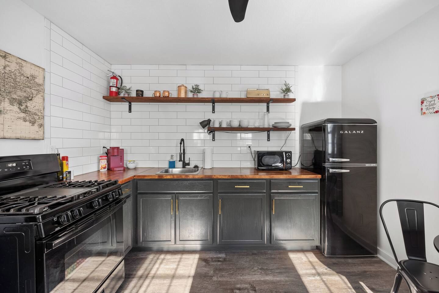A modern kitchen with a gas stove, microwave, and Galanz refrigerator. There are wooden shelves above the sink and a map on the wall.