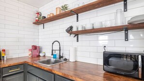 Una cocina con baldosas blancas tipo metro, estantes de madera, un microondas negro y un lavamanos de acero inoxidable.