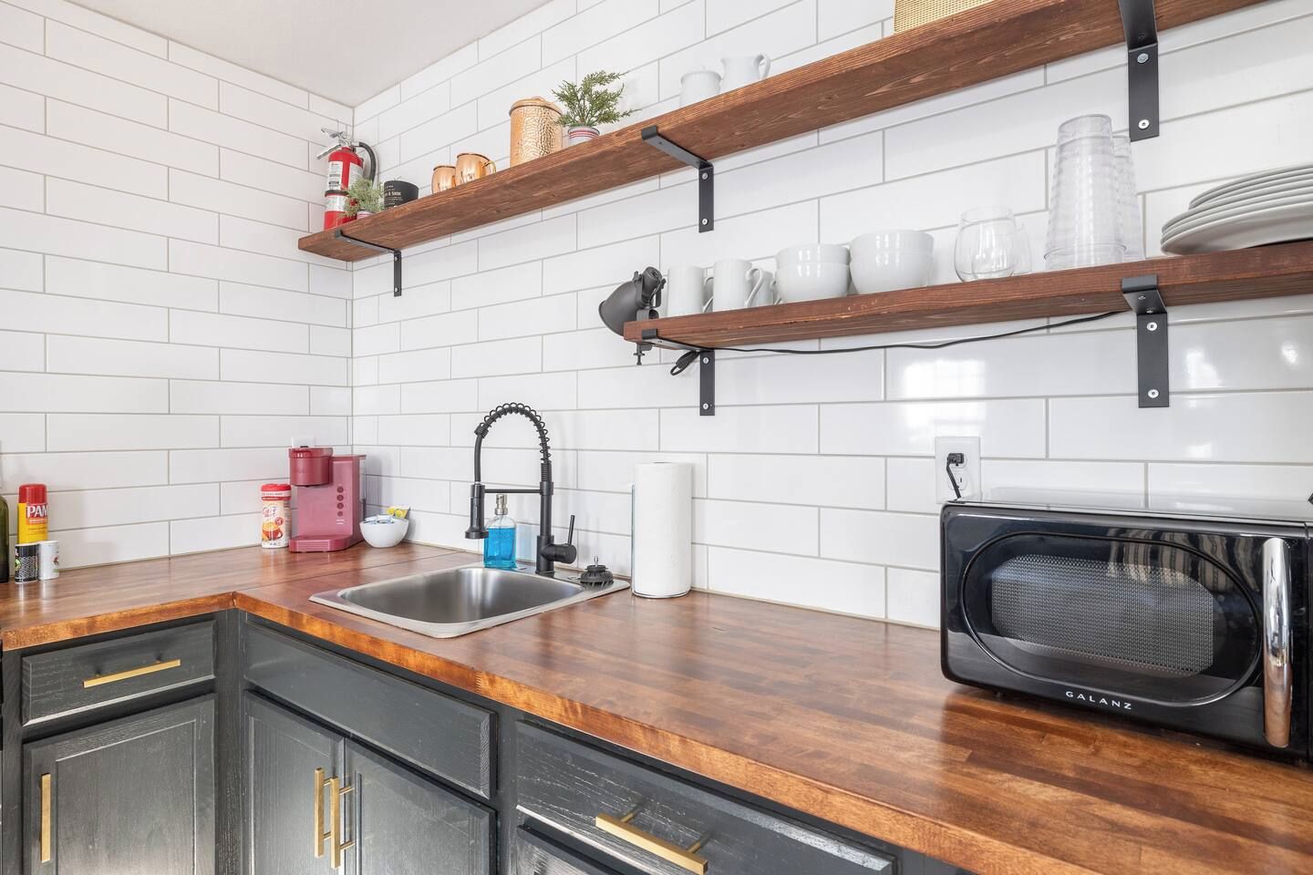 Een keuken met een roestvrijstalen spoelbak, houten planken, een zwarte magnetron en een witte betegelde achterwand.