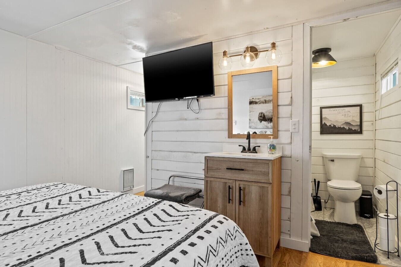 Ein Zimmer mit einem Bett, einem Nachttisch mit Spiegel, einer Toilette und einem an der Wand befestigten Fernseher.