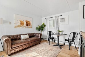 Una sala de estar con un sofá de cuero marrón, una mesa de comedor con dos sillas, una planta en maceta y un cuadro de ganado en la pared.