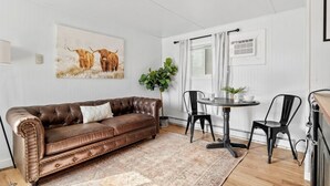 A living room with a brown leather sofa, a round table with chairs, a potted plant, and a painting of cattle on the wall. - Cabin #11 - Spearfish Orchard Creek Cottages (Spearfish)