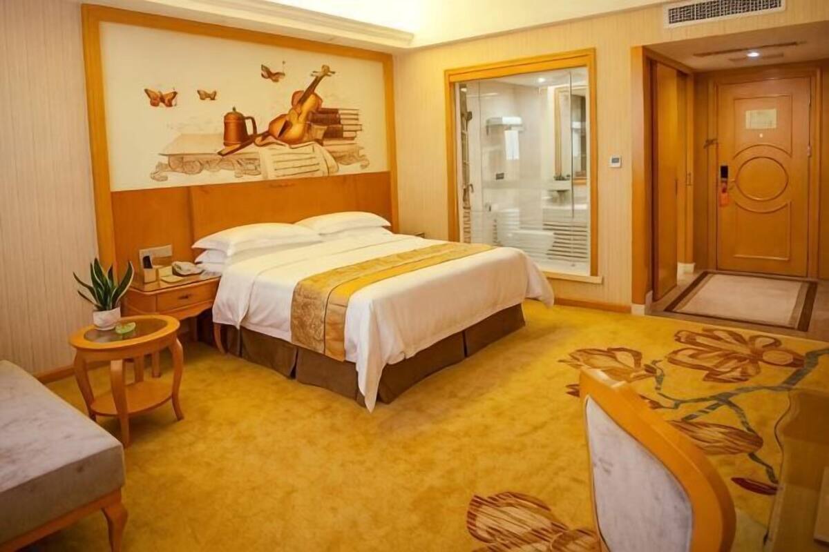 Ett hotellrum med en säng, ett nattduksbord, en stol, ett litet bord med en växt och en dörr som leder till ett badrum.
