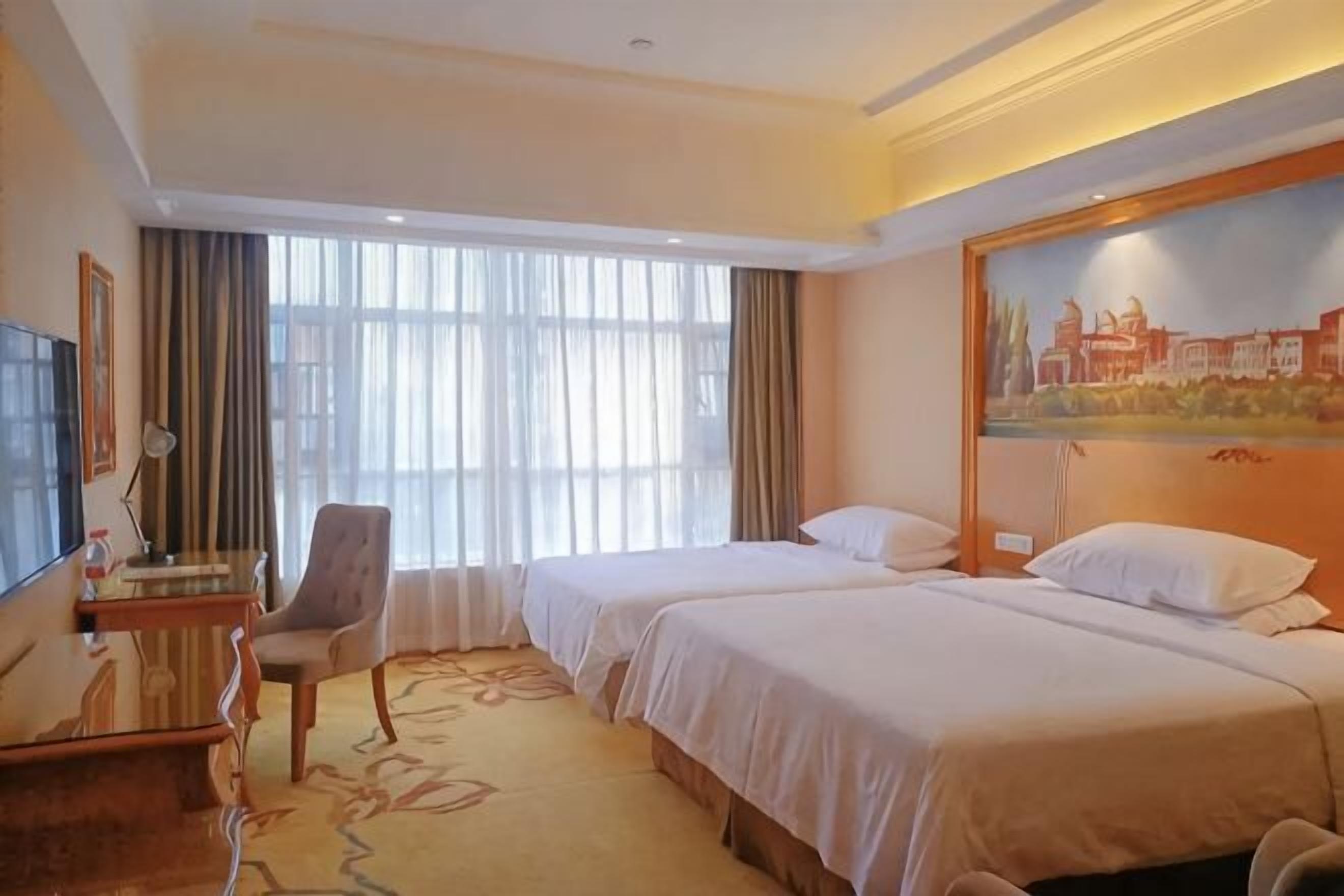 A hotel room with two beds, a desk with a chair, a TV, and a painting on the wall.