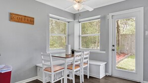 Een eethoek met een witte tafel en stoelen, een plafondventilator en een raam met uitzicht op de achtertuin.