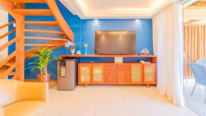 A modern living room with a wooden staircase, a flat-screen TV, a small refrigerator, a sofa, and a potted plant. - IT32 Triplex 4 Suítes a 100m da Praia (Camaçari)