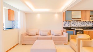 A modern living area with a sofa, ottoman, and chairs, a kitchen with a stove and microwave, and a window with curtains. - IT32 Triplex 4 Suítes a 100m da Praia (Camaçari)