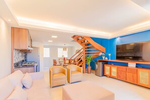 Un salon moderne comprenant un canapé, deux fauteuils, un téléviseur, un escalier en bois et un coin cuisine.
