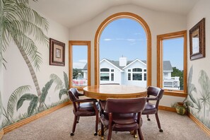A dining area with a round table and four chairs, large windows with wooden frames, and a view of a white house with a balcony.