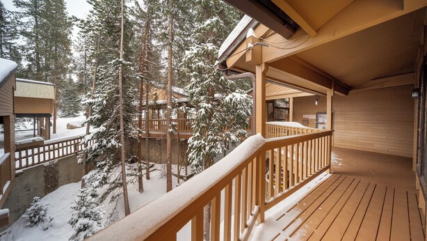 A wooden deck with a railing, surrounded by snow-covered trees and a building with a wooden exterior.