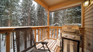 A wooden deck with a grill and a chair, overlooking a snowy forest.
