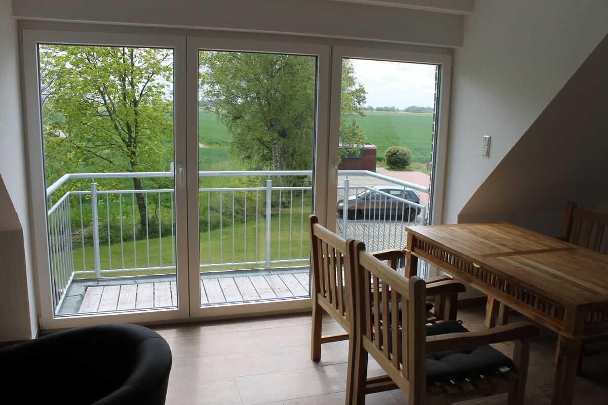 A balcony with a wooden table and chairs, a view of a green landscape, and a white railing.