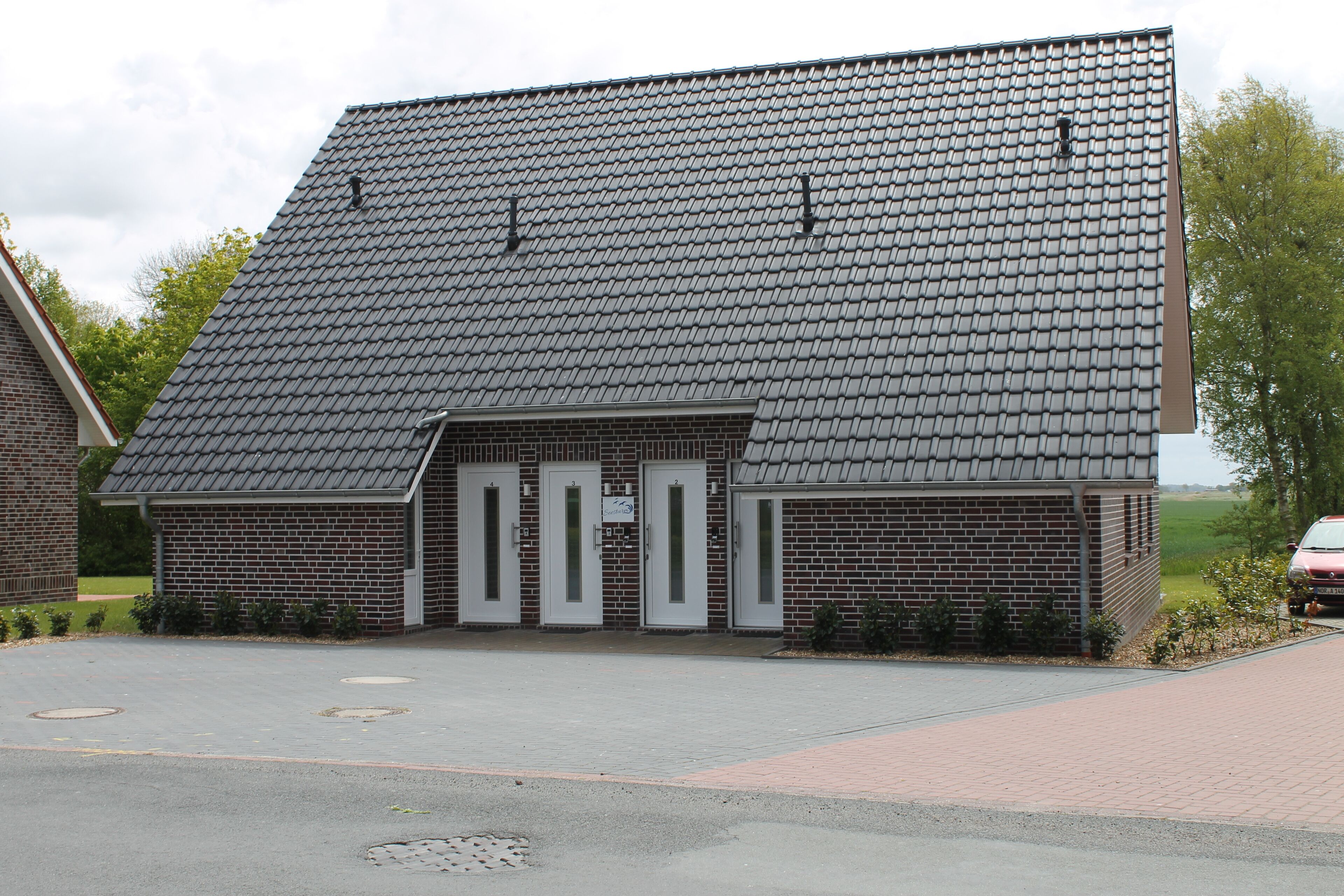 Ein Gebäude aus Backsteinen mit einem geneigten Dach, drei Eingängen und einer gepflasterten Auffahrt.