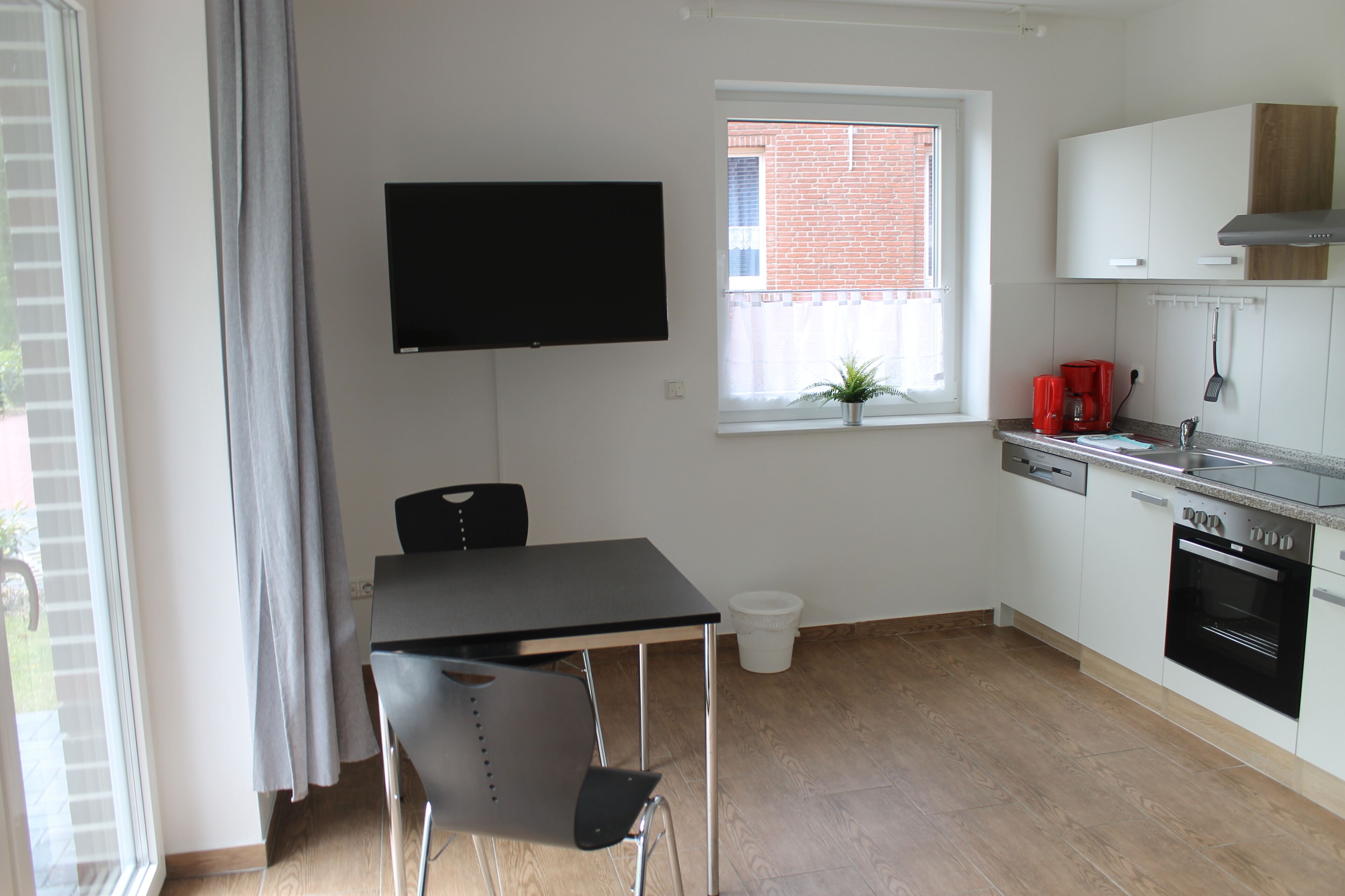 Cocina compacta con mesa de comedor, televisor montado en la pared, ventana con planta y horno empotrado.