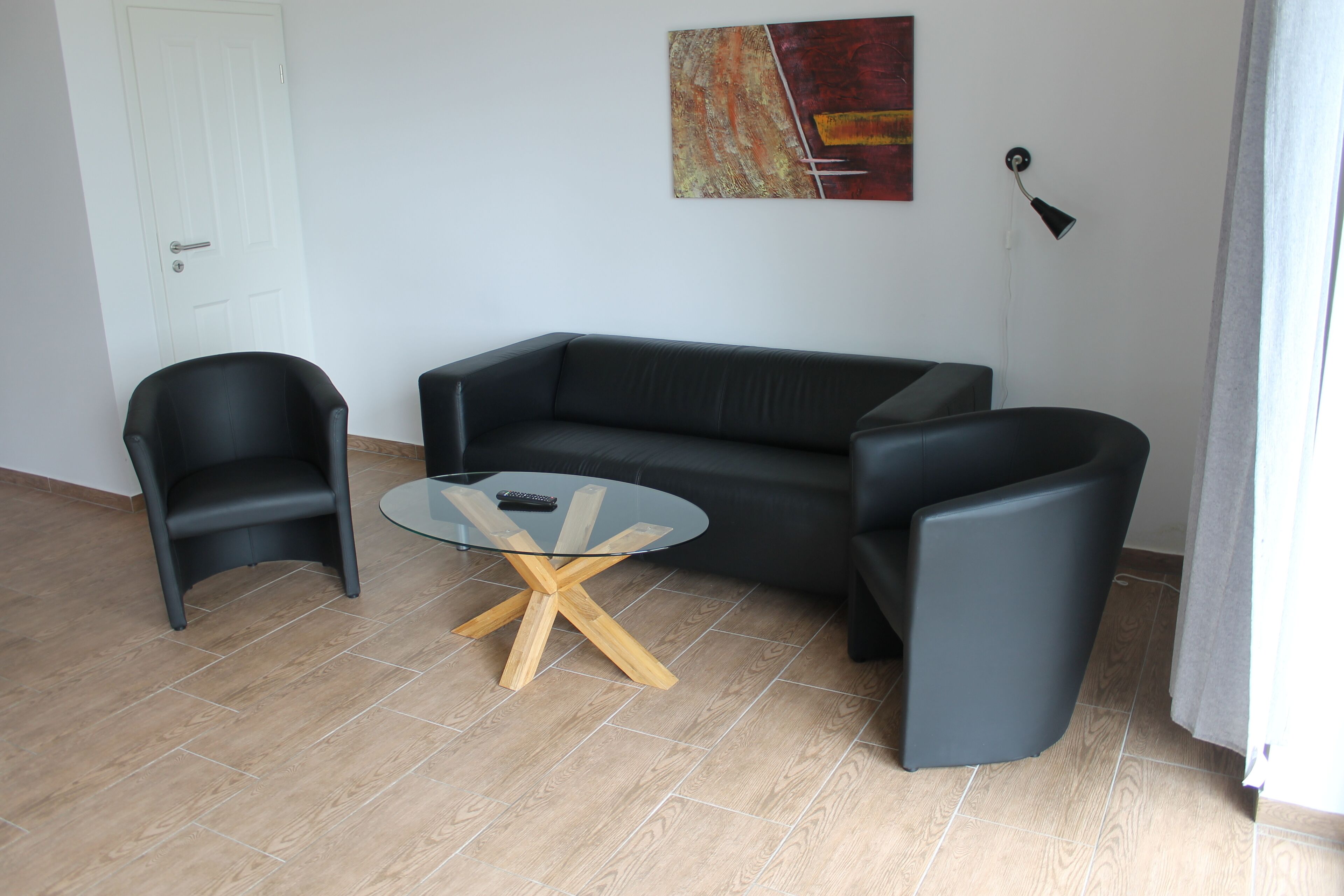 Een moderne woonkamer met een zwarte leren bank, een glazen salontafel met houten poten en twee zwarte fauteuils.
