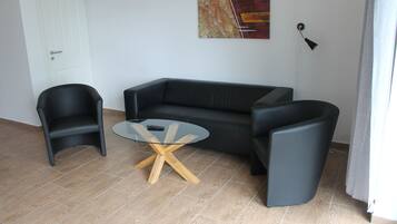 Een moderne woonkamer met een zwarte leren bank, een glazen salontafel met houten poten en twee zwarte fauteuils.
